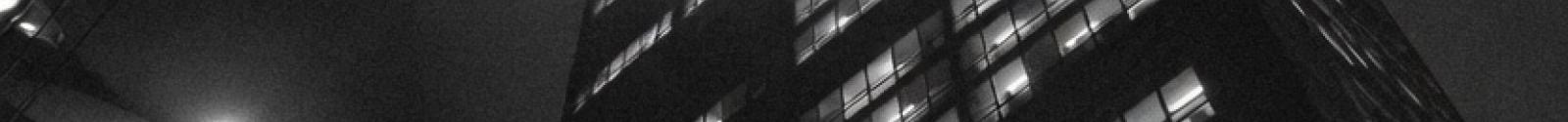 Black and white photo of tall office buildings lit up at night, shot from ground level looking up