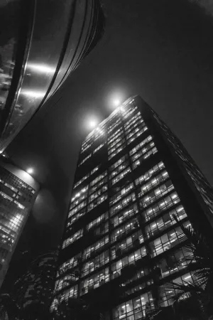 Black and white photo of tall office buildings lit up at night, shot from ground level looking up